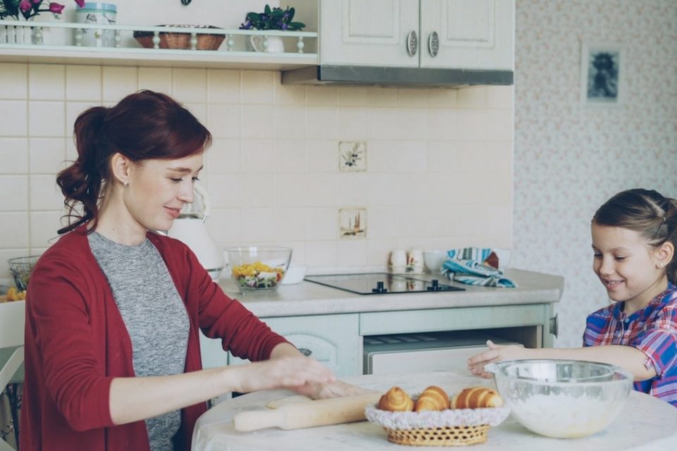Young mother rolling dough and talking to her little cute daughter while cooking in the kitchen on weekend. Family, food, home and people concept