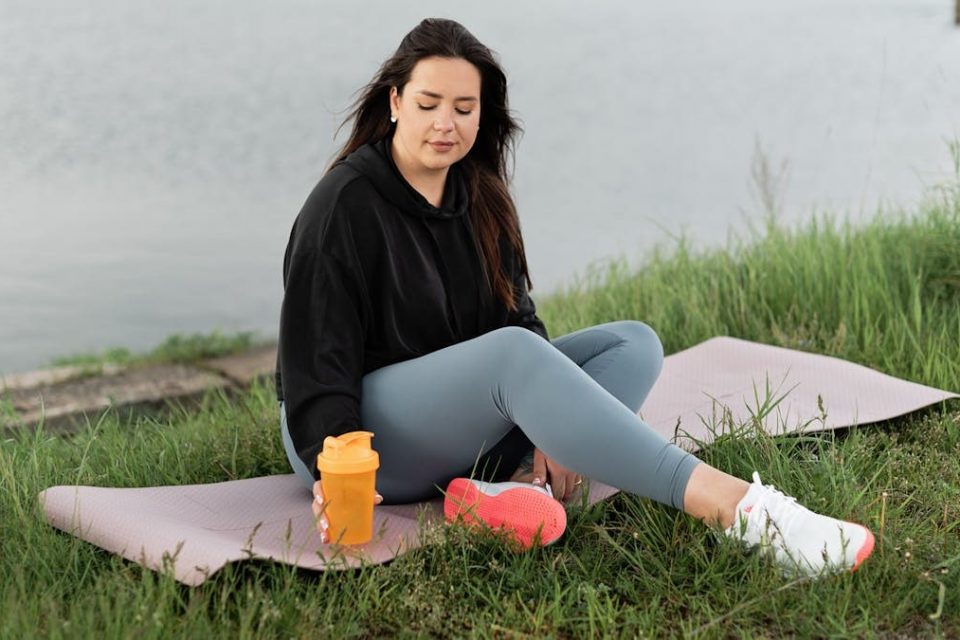 Woman enjoying a peaceful moment by the lake with a yoga mat and water bottle, promoting wellness.